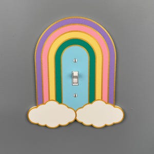 May include: A rainbow-shaped light switch cover featuring concentric arches in purple, pink, yellow, and green, with a light blue centre panel. The design includes two white clouds at the base and is secured by two silver screws on the switch.