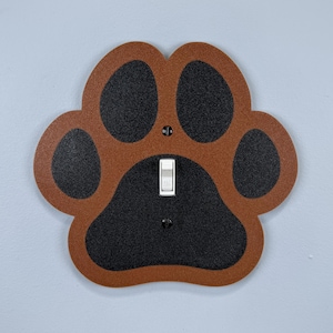 May include: A decorative light switch cover shaped like a dog paw print. The paw print is brown with black pads and a white light switch in the center. The cover is mounted on a light blue wall.