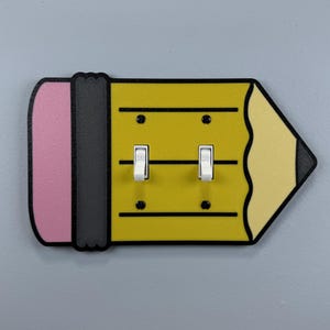 May include: A decorative light switch cover shaped like a yellow pencil with a pink eraser and black accents. It features two white toggle switches and is mounted on a gray wall. The pencil design adds a playful touch to any room.