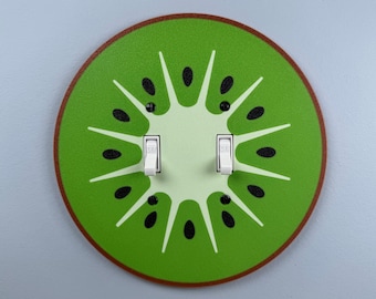 May include: A round light switch cover featuring a kiwi fruit design. The cover is green with white and black accents, and a brown border. Two white light switches are in the center.