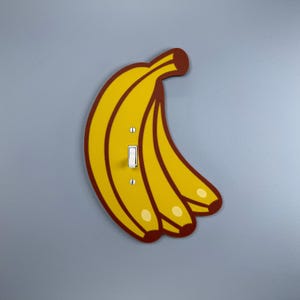 May include: A light switch cover with a banana design. The cover is yellow with brown outlines and stems, and features a white toggle switch. It is mounted on a grey wall. The banana design adds a playful touch to any room.
