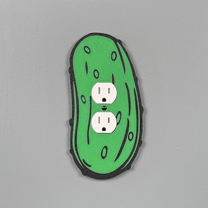 May include: A green and black decorative electrical outlet cover shaped like a pickle. The cover has two white electrical outlets and is mounted on a gray wall. The pickle design includes black outlines and details.