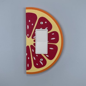 May include: A decorative light switch cover shaped like a half grapefruit slice. The design features a white light switch at the centre, set against a red and yellow fruit pattern. The cover is mounted on a light blue wall.