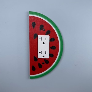 May include: A decorative wall outlet cover shaped like a watermelon slice. The cover features a red interior with black seed details, a white outlet, and a green rind. The outlet cover is mounted on a light blue wall.