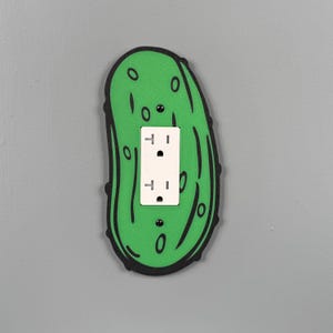 May include: A green pickle-shaped light switch cover with a black outline and white electrical outlet. The cover is mounted on a gray wall, adding a touch of humor to home decor.