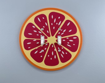 May include: A decorative light switch cover featuring a grapefruit slice design. The cover has a red and yellow color scheme with white accents and two white light switches. The cover is mounted on a gray wall.