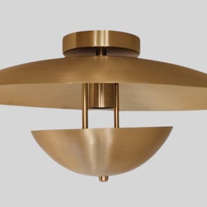 May include: A contemporary ceiling light in a brass finish. It has a wide, disc-shaped shade and a smaller, bowl-like element hanging beneath. The design is minimalist and stylish, suitable for different interior designs.
