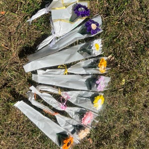 Op de afbeelding: Een verzameling kleurrijke, enkelvoudige kunstbloemen, elk verpakt in transparant cellofaan en wit papier. De bloemen zijn er in verschillende kleuren: paars, wit, geel, roze en oranje, gerangschikt op groen gras.