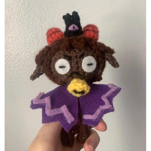 May include: A handmade crochet doll with a whimsical design. The doll features a brown head with white eyes, a yellow beak, and a purple cape with zigzag edges. It has red ear-like accents and a black crown.