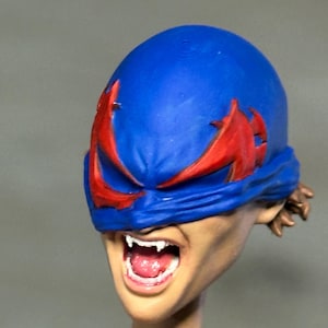 May include: A sculpted head of a figure featuring a bright blue mask with jagged red eye cutouts. The figure has a light skin tone, an open mouth revealing white teeth and a pink tongue, and brown hair visible at the back of the head.