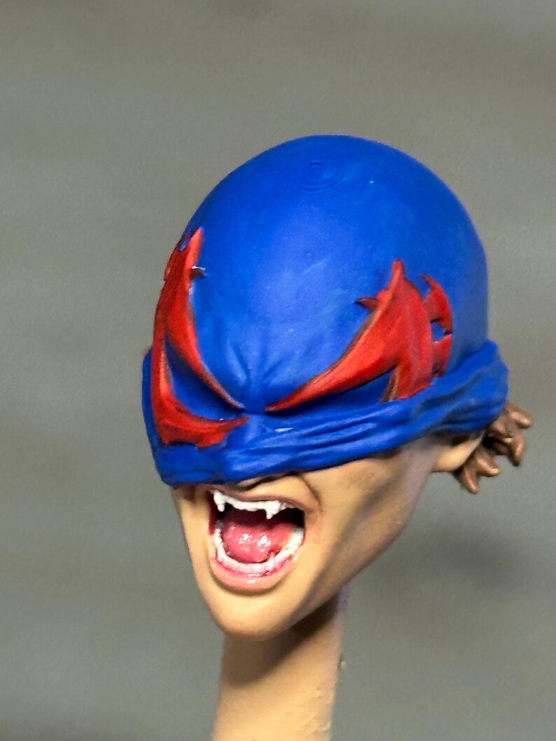 May include: A sculpted head of a figure featuring a bright blue mask with jagged red eye cutouts. The figure has a light skin tone, an open mouth revealing white teeth and a pink tongue, and brown hair visible at the back of the head.