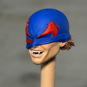 May include: A sculpted action figure head featuring a blue mask with red wing-like accents covering the eyes. The face has a tan skin tone, a wide grin revealing sharp white fangs, and brown hair visible at the back of the head, mounted on a post.