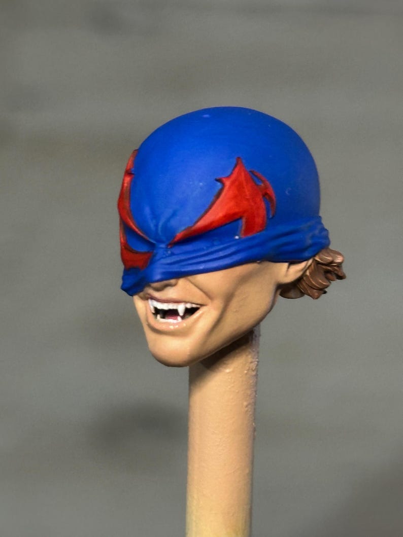 May include: A sculpted action figure head featuring a blue mask with red wing-like accents covering the eyes. The face has a tan skin tone, a wide grin revealing sharp white fangs, and brown hair visible at the back of the head, mounted on a post.
