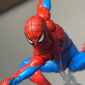 May include: A red and blue Spider-Man action figure in a dynamic pose. The figure features a detailed red mask with white eye lenses, a red and blue suit with web detailing, and a black spider emblem on the chest. The figure is supported by a clear stand.