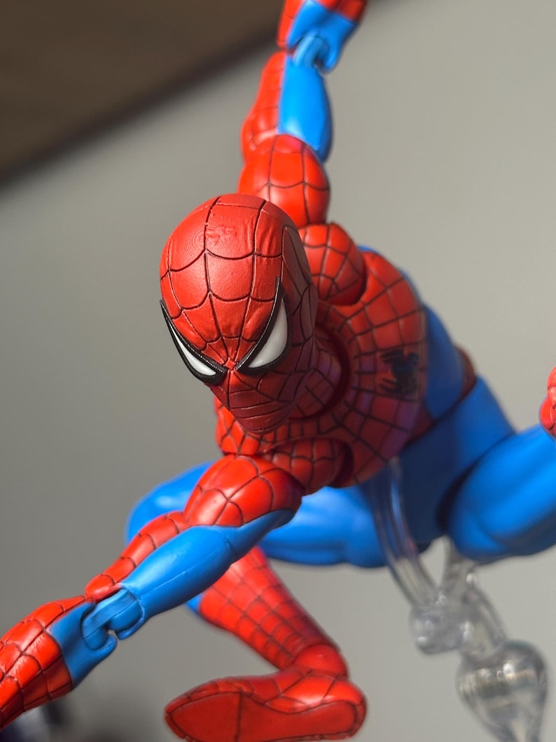 May include: A red and blue Spider-Man action figure in a dynamic pose. The figure features a detailed red mask with white eye lenses, a red and blue suit with web detailing, and a black spider emblem on the chest. The figure is supported by a clear stand.