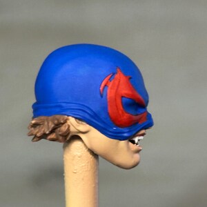 May include: A sculpted action figure head featuring a bright blue mask with a prominent red emblem over the eyes. The figure has light skin, visible brown hair at the base of the mask, and an open mouth revealing white teeth in a snarling expression.