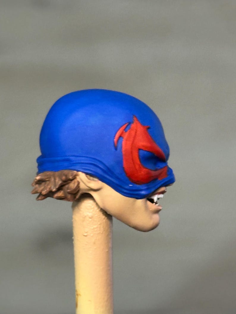 May include: A sculpted action figure head featuring a bright blue mask with a prominent red emblem over the eyes. The figure has light skin, visible brown hair at the base of the mask, and an open mouth revealing white teeth in a snarling expression.