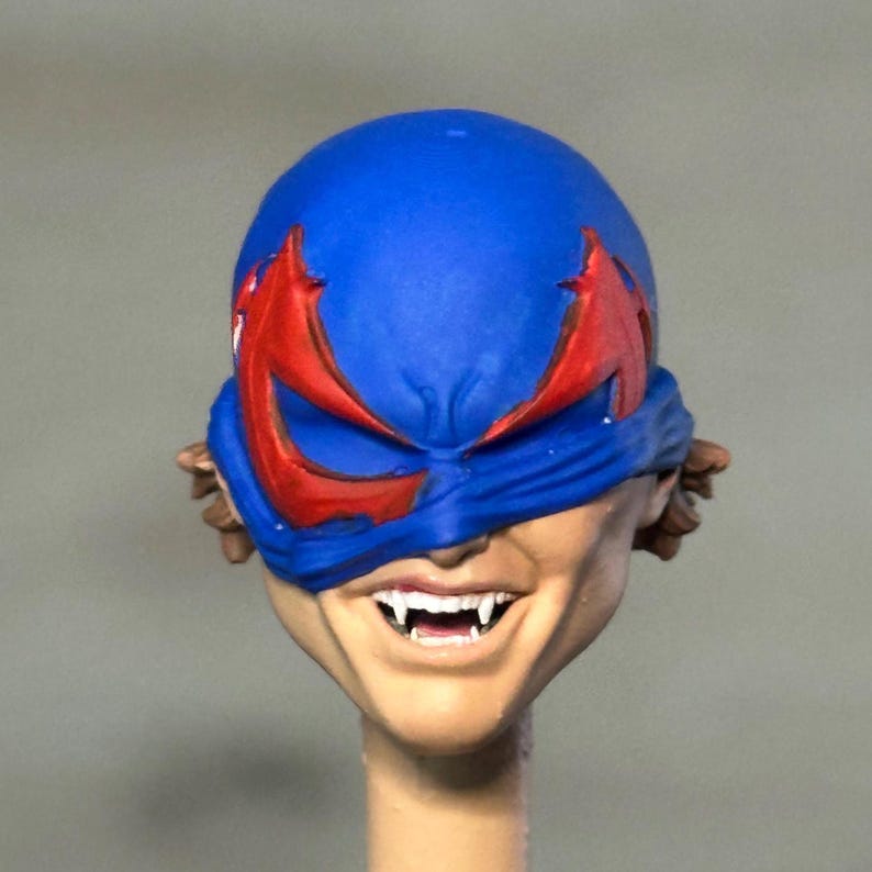 May include: A sculpted figurine head featuring a blue mask with jagged red eye cutouts. The face has a light skin tone, a wide grin, and visible white fangs. Brown hair is partially visible at the sides, and the head is mounted on a cylindrical neck post.