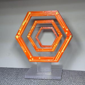 May include: An orange and yellow geometric sculpture featuring nested hexagonal shapes. The artwork has a circuit board design with yellow lines and dots. It is mounted on a clear, rectangular base.