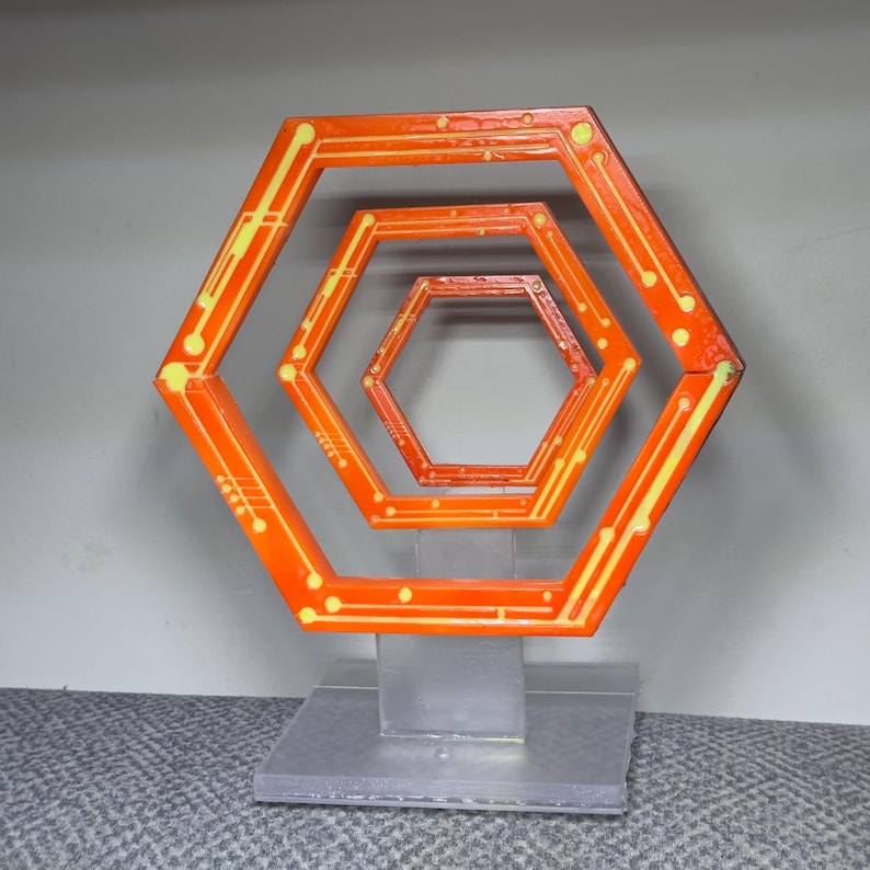 May include: An orange and yellow geometric sculpture featuring nested hexagonal shapes. The artwork has a circuit board design with yellow lines and dots. It is mounted on a clear, rectangular base.