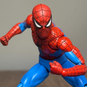 May include: A red and blue Spider-Man action figure in a dynamic pose. The figure has a red mask with white eye lenses, a red torso and arms with a web pattern, and blue pants. The figure is posed with clenched fists.