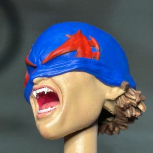May include: A sculpted action figure head featuring a blue mask with red accents covering the eyes. The face has a light skin tone, an open mouth revealing white teeth, and brown hair visible at the back. The head is mounted on a cylindrical post.