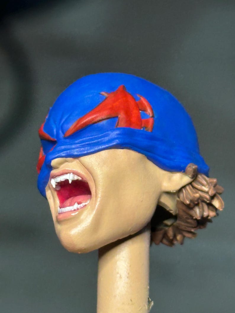 May include: A sculpted action figure head featuring a blue mask with red accents covering the eyes. The face has a light skin tone, an open mouth revealing white teeth, and brown hair visible at the back. The head is mounted on a cylindrical post.