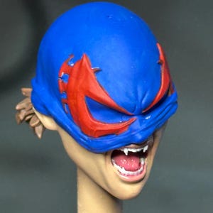 May include: A sculpted figurine head featuring a bright blue mask with sharp red eye accents. The face has an open mouth revealing white teeth and fangs, with light brown hair visible at the back. The skin tone is a light beige, and the neck is cylindrical.