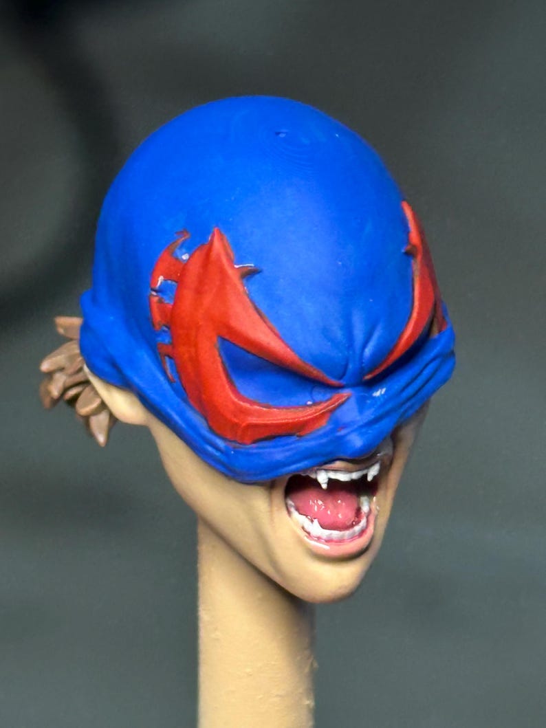 May include: A sculpted figurine head featuring a bright blue mask with sharp red eye accents. The face has an open mouth revealing white teeth and fangs, with light brown hair visible at the back. The skin tone is a light beige, and the neck is cylindrical.