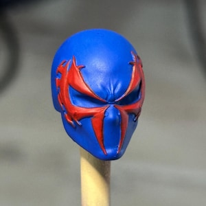 May include: A blue sculpted head accessory featuring a prominent red spider-like emblem that extends across the eye area. The surface is smooth with molded details around the brow and nose, showcasing a vibrant color contrast between the blue and red.