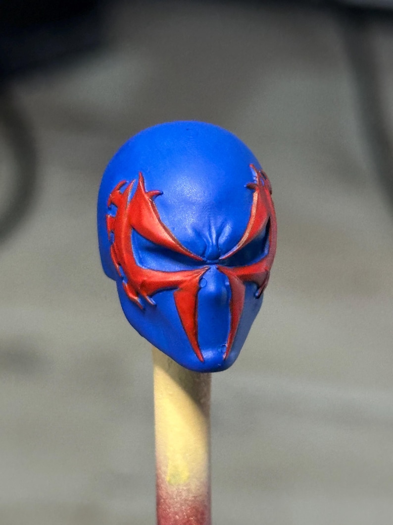 May include: A blue sculpted head accessory featuring a prominent red spider-like emblem that extends across the eye area. The surface is smooth with molded details around the brow and nose, showcasing a vibrant color contrast between the blue and red.