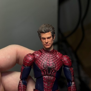Może przedstawiać: Szczeg&oacute;łowa figurka Spider-Mana, w czerwono-niebieskim kostiumie z pajęczyną. Figurka ma realistyczną rzeźbę głowy i jest trzymana przez dłoń. Kostium figurki ma teksturowany wygląd.