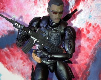 May include: A black action figure of a character wearing a tactical suit and sunglasses, holding a black rifle. The figure has a serious expression and a brown belt. A sword is strapped to the back. The background is a mix of red, white, and blue paint strokes.