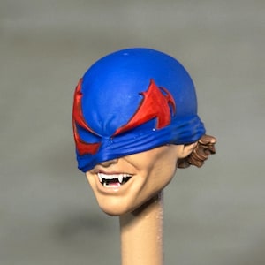 May include: A miniature sculpted head featuring a blue mask with red wing-like eye accents. The face has a light skin tone, a wide grin revealing sharp white fangs, and brown hair visible at the back. The head is mounted on a thin cylindrical post.