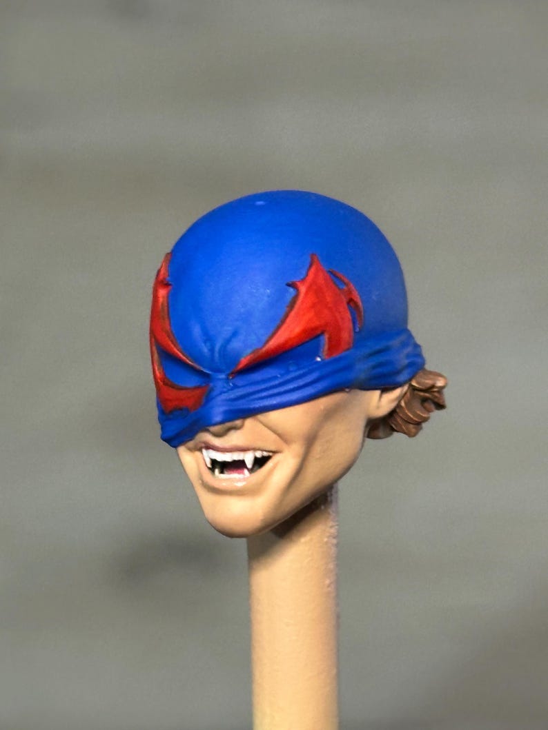 May include: A miniature sculpted head featuring a blue mask with red wing-like eye accents. The face has a light skin tone, a wide grin revealing sharp white fangs, and brown hair visible at the back. The head is mounted on a thin cylindrical post.