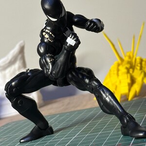 May include: A black action figure in a crouched fighting stance with white eye lenses and white tape accents on the hands and shoulder. The figure is positioned on a green cutting mat with visible white measurement markings in inches.