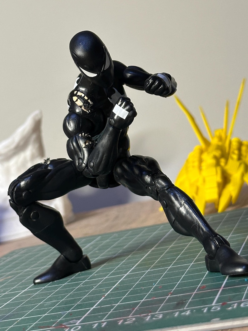 May include: A black action figure in a crouched fighting stance with white eye lenses and white tape accents on the hands and shoulder. The figure is positioned on a green cutting mat with visible white measurement markings in inches.