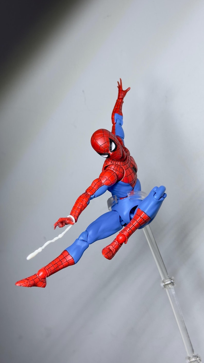 Puede incluir: Figura de acci&oacute;n de Spider-Man, rojo y azul, en pose din&aacute;mica, con un brazo levantado y una telara&ntilde;a que se extiende desde la mu&ntilde;eca. La figura presenta un patr&oacute;n texturizado y est&aacute; suspendida sobre un fondo neutro.