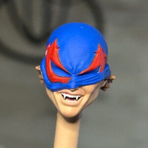 May include: A sculpted action figure head featuring a blue mask with jagged red eye cutouts. The face has a light skin tone, a wide grin, and visible sharp white fangs. The head is mounted on a thin, cylindrical beige support post.