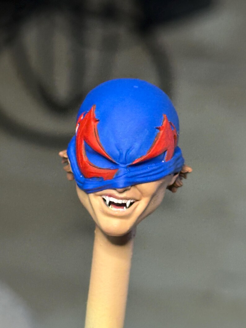 May include: A sculpted action figure head featuring a blue mask with jagged red eye cutouts. The face has a light skin tone, a wide grin, and visible sharp white fangs. The head is mounted on a thin, cylindrical beige support post.