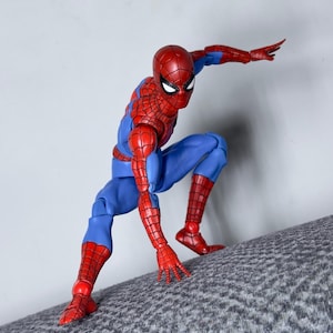 Puede incluir: Figura de acci&oacute;n de Spider-Man en una pose din&aacute;mica, vestida con un traje rojo y azul. La figura est&aacute; agachada sobre una superficie texturizada, con un brazo extendido. La m&aacute;scara de la figura tiene lentes blancos.