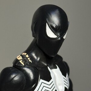 May include: A black action figure with a smooth, matte finish head and large, angular white eyes. The figure features a white spider-like emblem on the chest and shows visible wear with chipped paint revealing a tan base on the shoulder area.