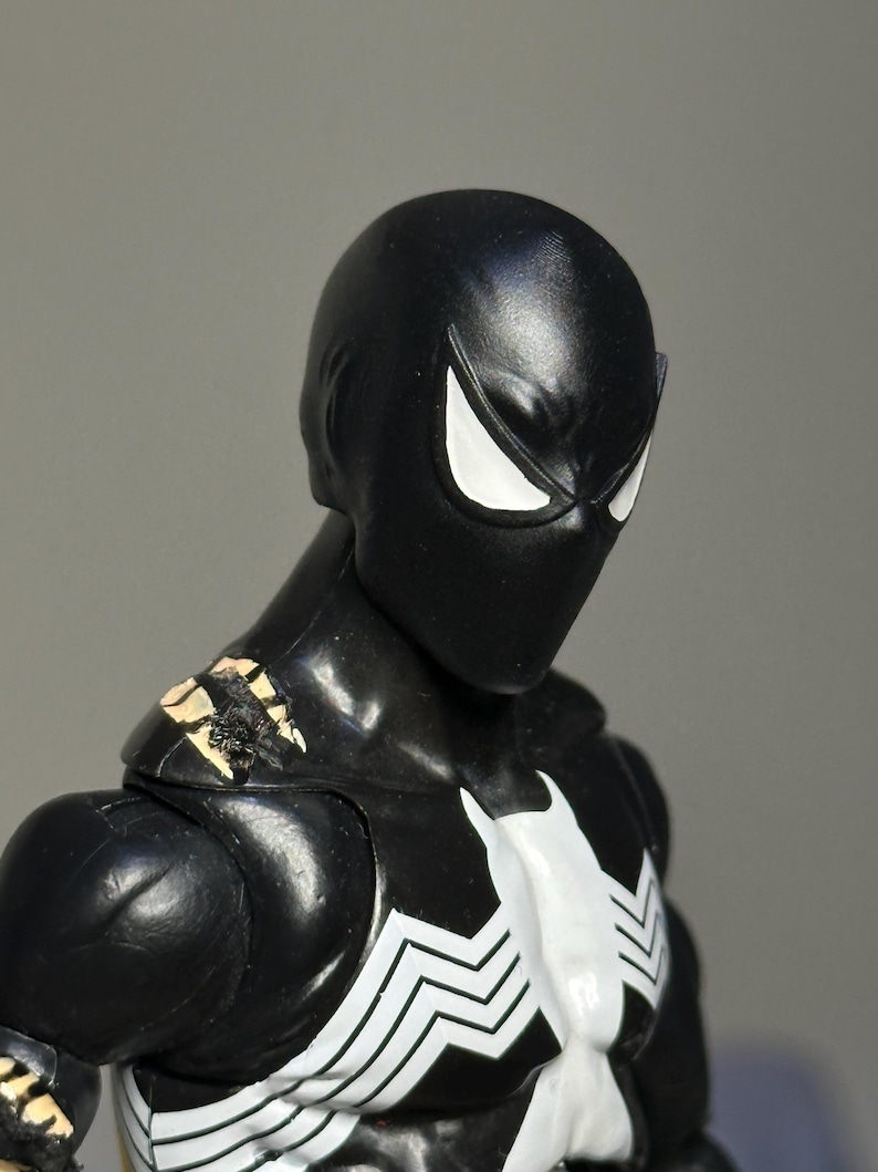 May include: A black action figure with a smooth, matte finish head and large, angular white eyes. The figure features a white spider-like emblem on the chest and shows visible wear with chipped paint revealing a tan base on the shoulder area.
