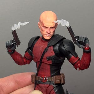 以下が含まれることがあります： 傷跡のある顔をしたハゲのフィギュアで、赤と黒のタクティカルスーツを着用しています。フィギュアは2丁の黒いピストルを構えており、銃口からは半透明の白い煙のエフェクトが出ています。茶色のユーティリティベルトと円形のバックルが特徴です。
