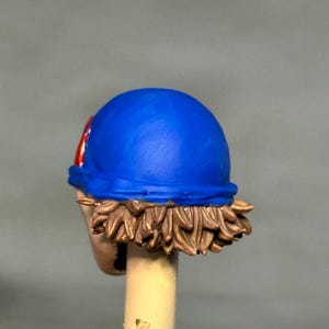 May include: A miniature figurine head featuring a bright blue cap with a red and white circular emblem on the side. The head has sculpted brown hair visible beneath the cap and is mounted on a light-colored cylindrical support rod.