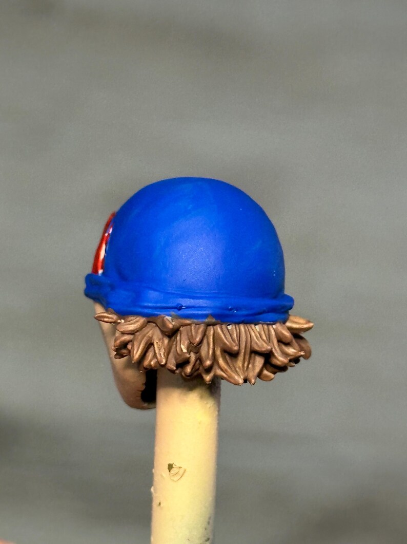 May include: A miniature figurine head featuring a bright blue cap with a red and white circular emblem on the side. The head has sculpted brown hair visible beneath the cap and is mounted on a light-colored cylindrical support rod.
