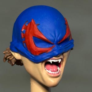 May include: A sculpted action figure head featuring a blue mask with red eye markings. The face has a light skin tone, an open mouth revealing white fangs and a pink tongue, and brown hair visible at the back of the head under the mask.
