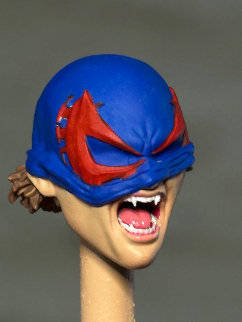 May include: A sculpted action figure head featuring a blue mask with red eye markings. The face has a light skin tone, an open mouth revealing white fangs and a pink tongue, and brown hair visible at the back of the head under the mask.