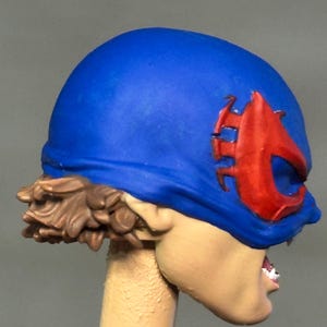 May include: A sculpted figurine head featuring a bright blue mask with a prominent red emblem over the eye area. The figure has short, textured brown hair visible at the back and a light skin tone, with a partially visible mouth showing white teeth.