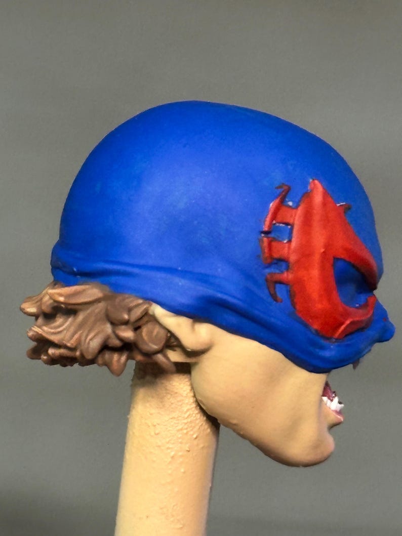 May include: A sculpted figurine head featuring a bright blue mask with a prominent red emblem over the eye area. The figure has short, textured brown hair visible at the back and a light skin tone, with a partially visible mouth showing white teeth.
